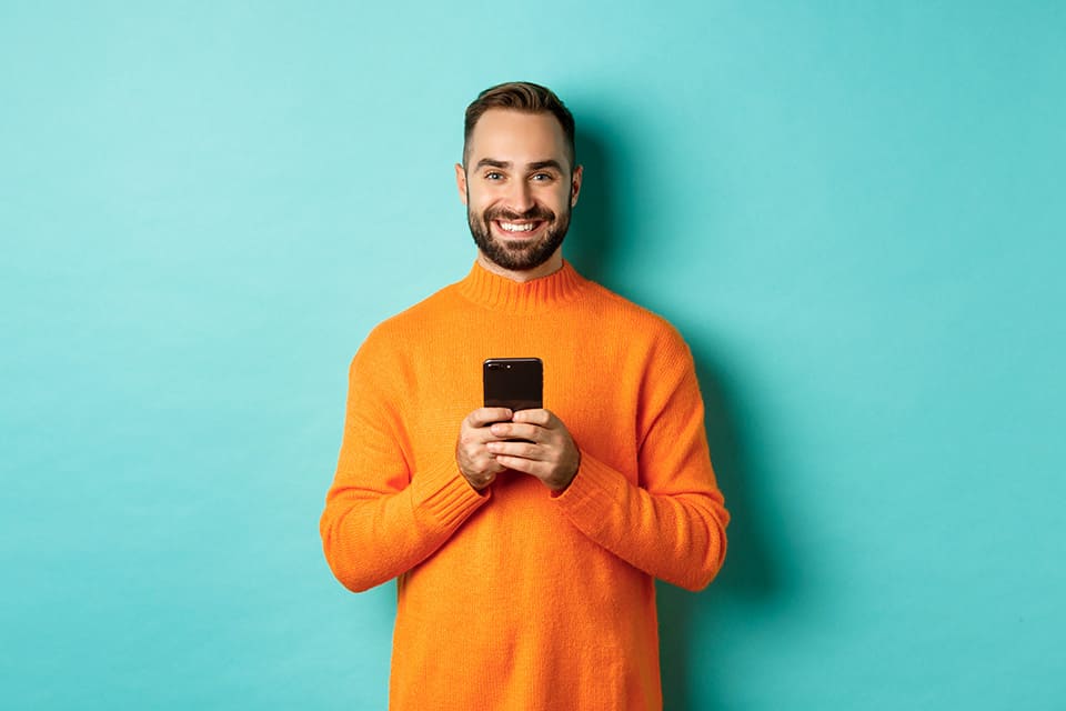 Homme souriant en pull orange tenant un smartphone, sur fond bleu turquoise.
