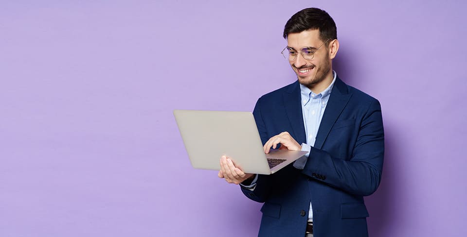Homme souriant en costume tenant un ordinateur portable sur un fond violet uni.